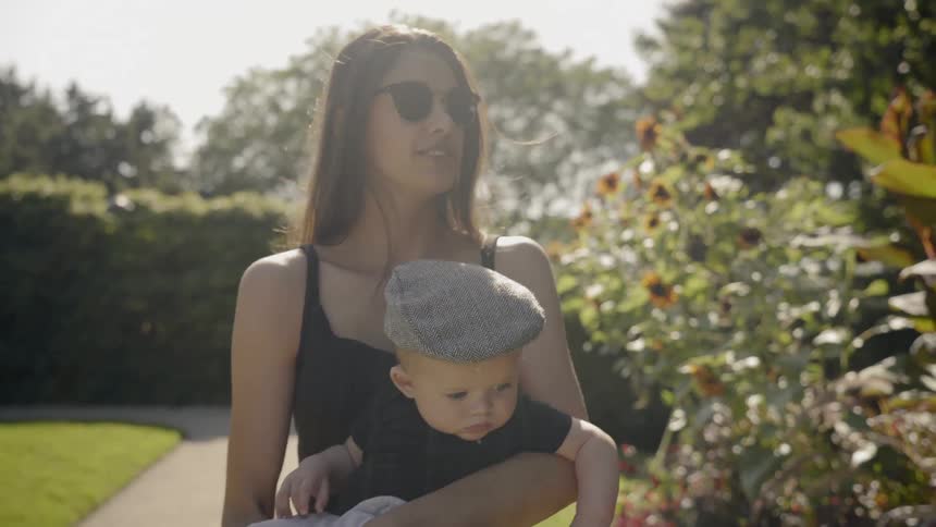 A woman holds her infant son as they walk down a sidewalk on a sunny day