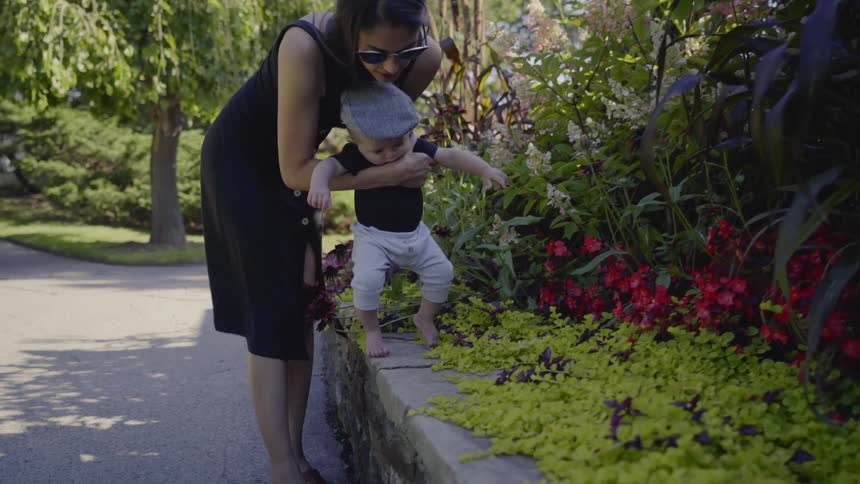 A woman helps her infant son try to walk along a garden ledge