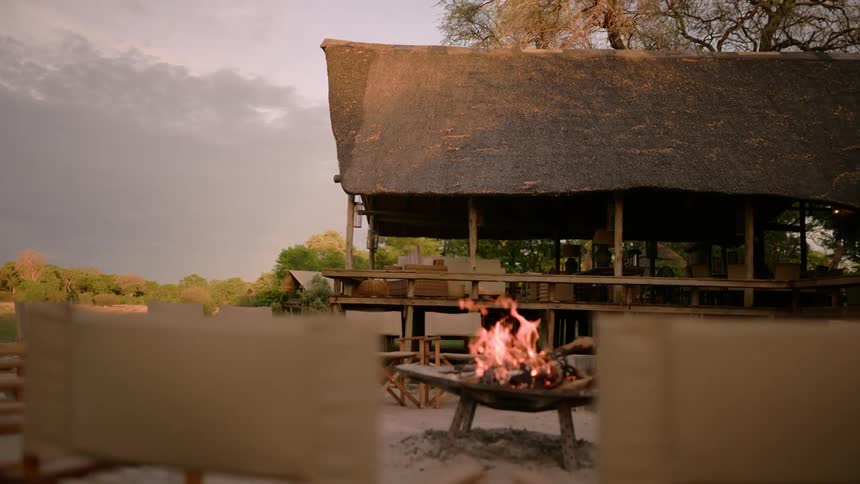 A fire pit surrounded by chairs and a thatched roof pavilion