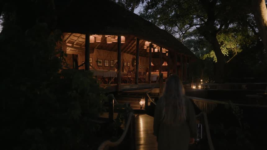 A woman walking along a dimly illuminated outdoor path towards a large wooden building