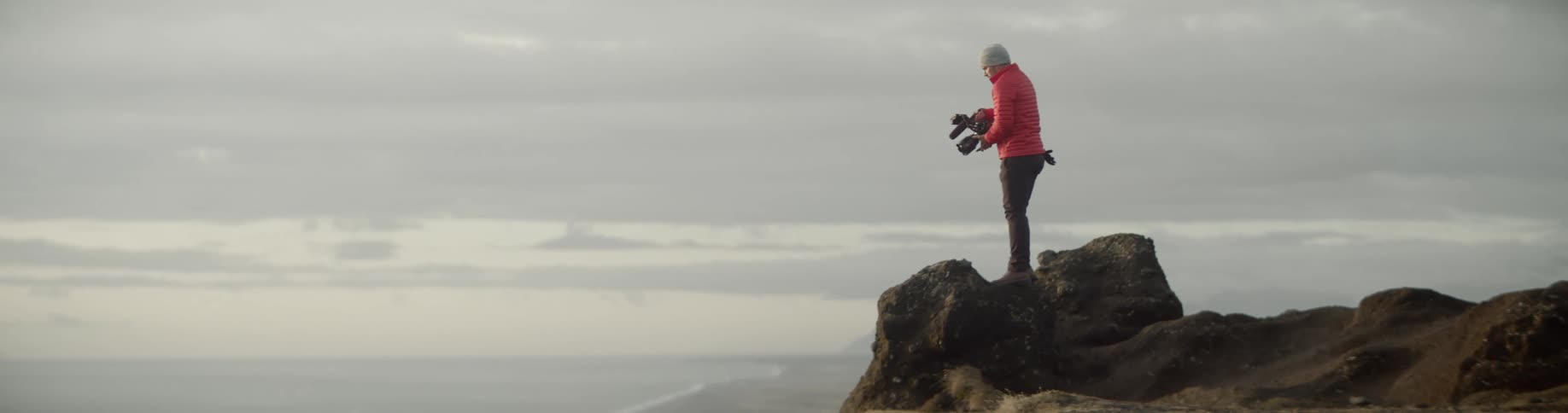 A man in a red winter coat stands on a cliff holding a camera and taking pictures of the ocean in front of him on a very cloudy day