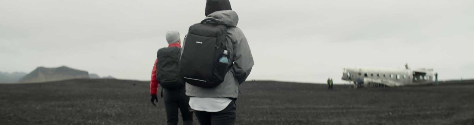 Two men wearing backpacks hike towards the abandoned and derelict remains of a passenger airplane