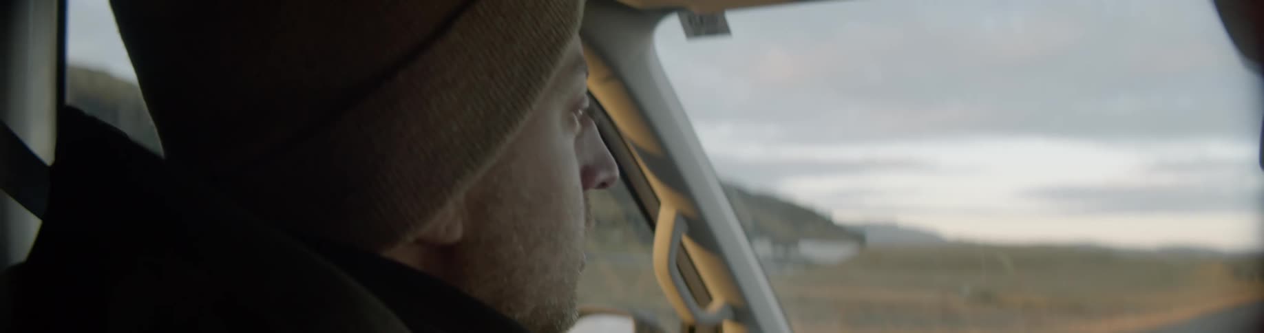 A man wearing a beanie drives along a remote road in a van