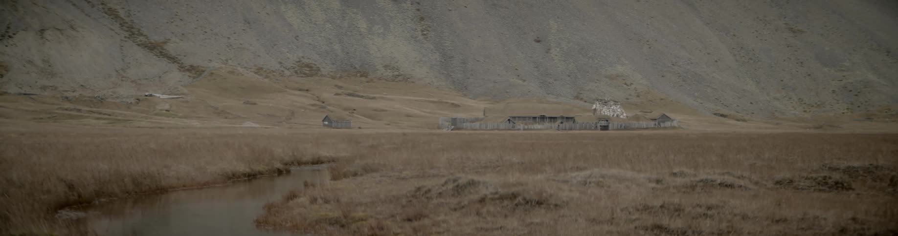 A small ranch house with a fence and outbuildings at the base of a hill