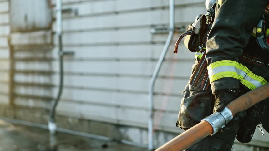 A firefighter in full protective gear stands outside while tugging a large fire hose