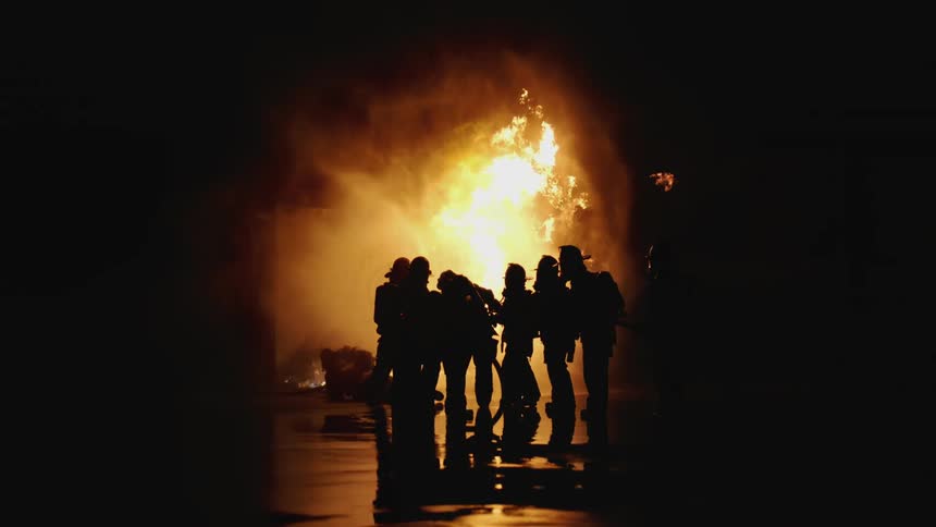 Firefighters stand before a flaring fire