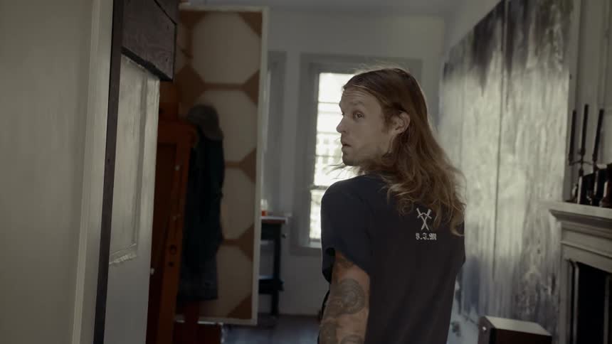 A young male with long hair and tattoos walks through an indoor hallway of a house towards a room with windows