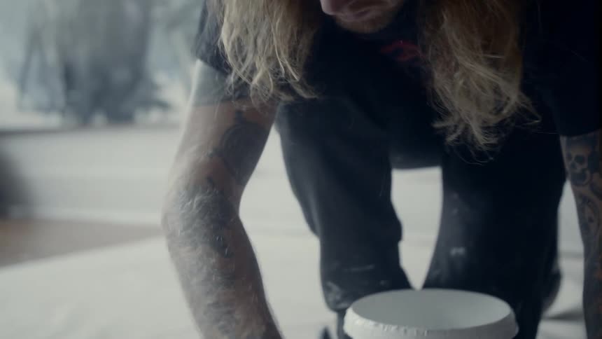 A man with long hair and a backwards baseball cap brushes white paint on a floor