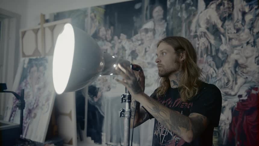Tattooed man with long hair moves a flood lamp inside painting studio