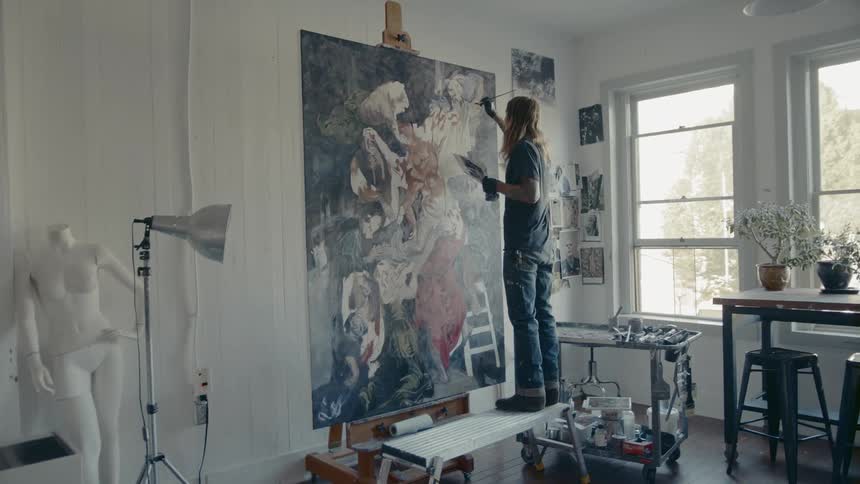 Man stands on bench to continue working on large painting near windows
