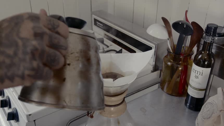Man with tattoos on his arm picks up a coffee pot from a stove and pours water into a filter
