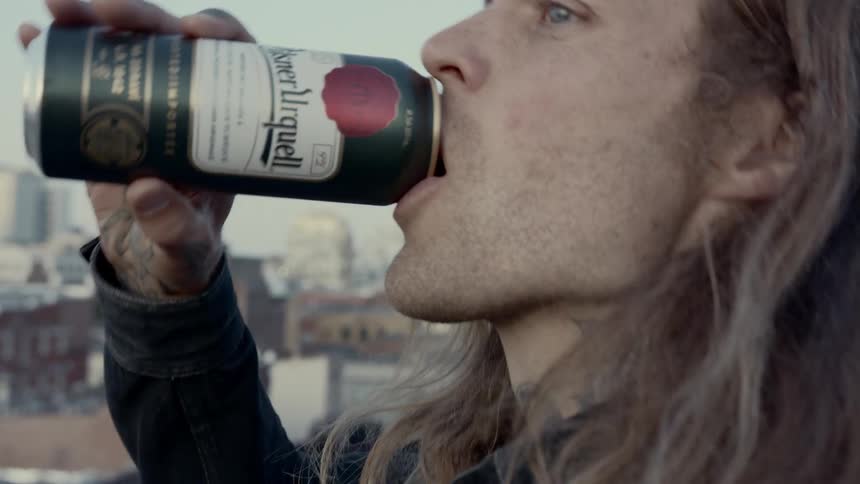 Man with long hair drinking beer on a balcony overlooking the city