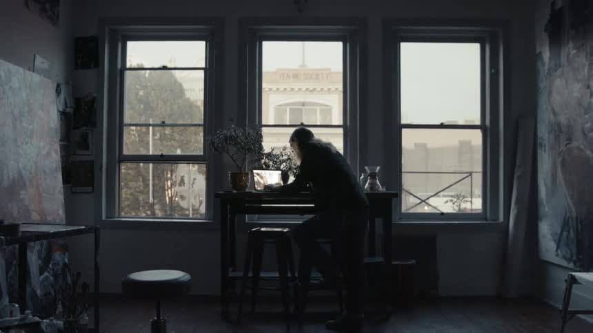 Long haired male painter uses a laptop leaning on desk in the workshop