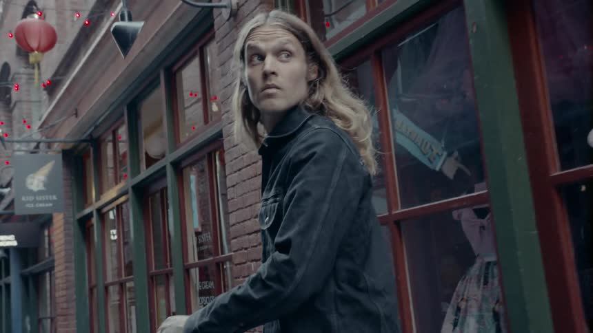 A man with long hair walks down an alley with shops on either side and keeps looking back over his shoulder