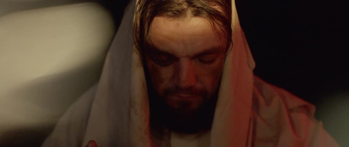 A man with a beard and wearing a white cloak wipes water on his face and looks upwards