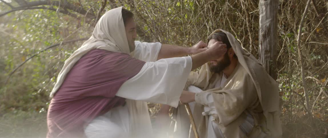 Man rubs the eyes of another man as they sit on the ground in the forest