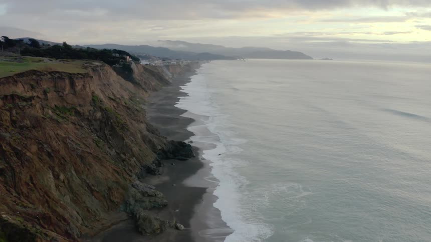 A coastline has a dark sand beach that is below a long row of cliffs
