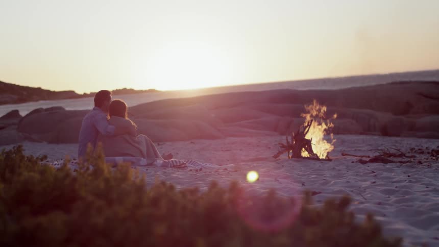 A man and woman sit cuddling by a campfire on a beach