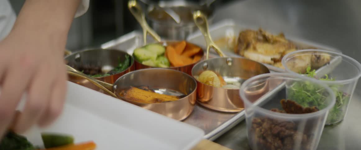 A chef in a commercial kitchen is preparing a plate of food from small pans of ingredients