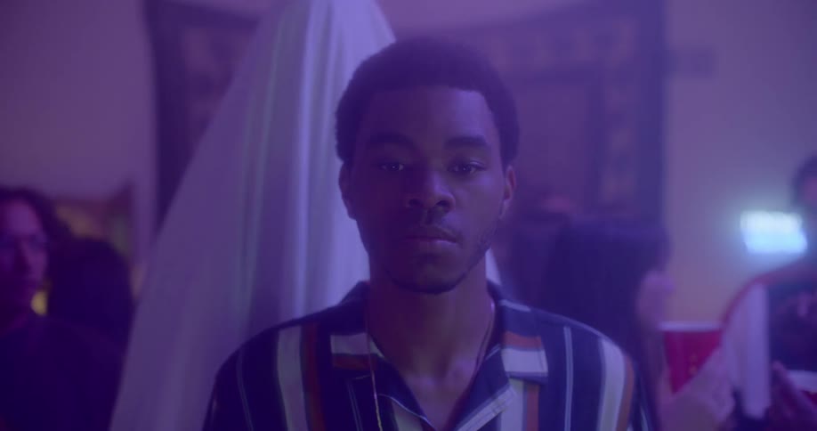A young black teenager stands alone in a crowded room of people dancing