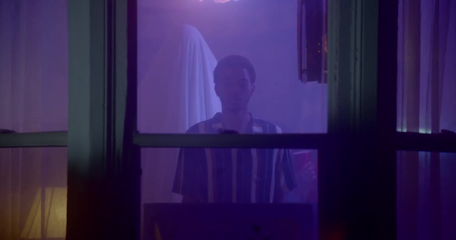 A young black male stands idly while staring out a window from a room with purple lighting