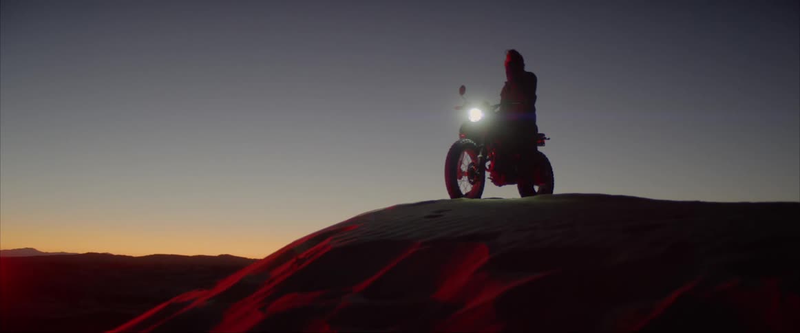 A man sits on a motorcycle atop a large hill at sunset and removes his helmet
