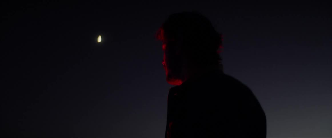 A man on a motorcycle taking his helmet off with the moon in the sky