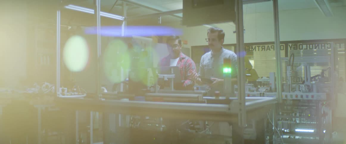 Two men talk as they control and watch a robotic arm