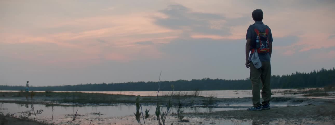 A man standing on the shore of a lake looking out over the water at sunset