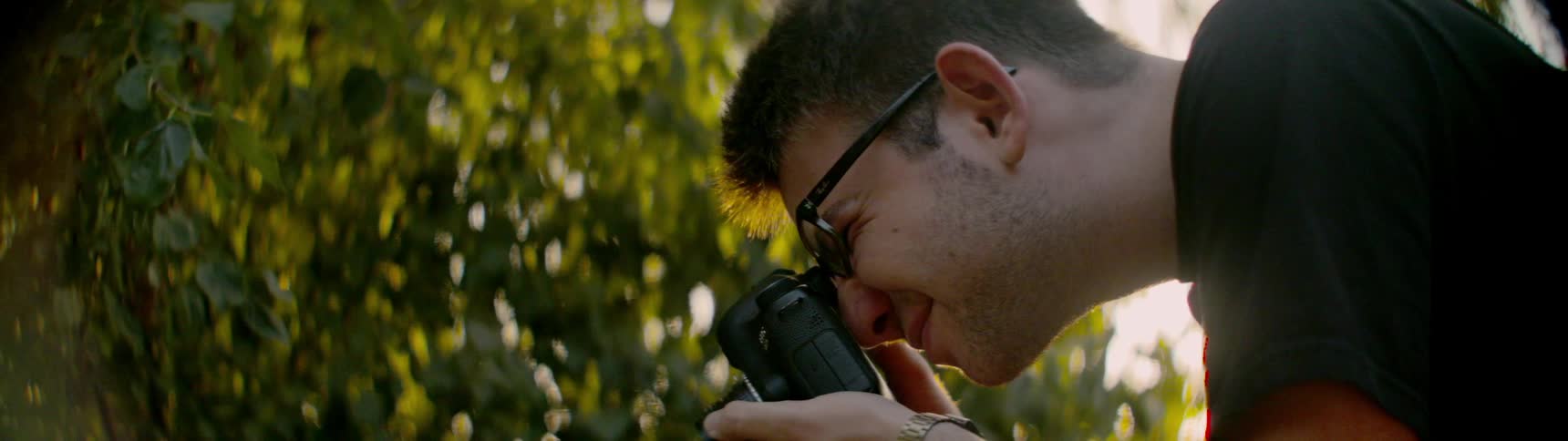 A man standing outdoors near a tree holds a camera and concentrates on taking a photograph