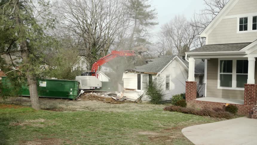 An excavator breaks through the roof of a small house as it demolishes it