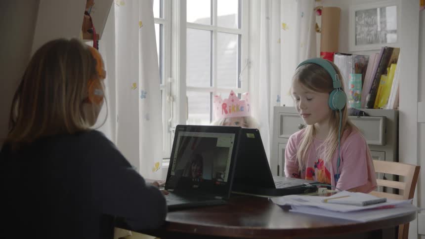 Two young white girls sit at a table listening to something on laptops