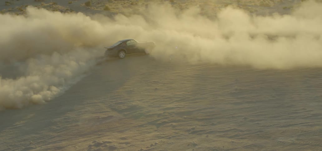 A porsche automobile is driving on a dirt covered clearing where is is making sharp turns and kicking up tons of dust