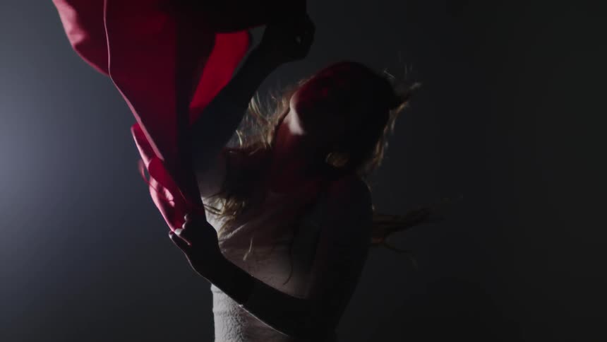 A woman holds a length of red material as a breeze blows it, and her hair, around in a dark room