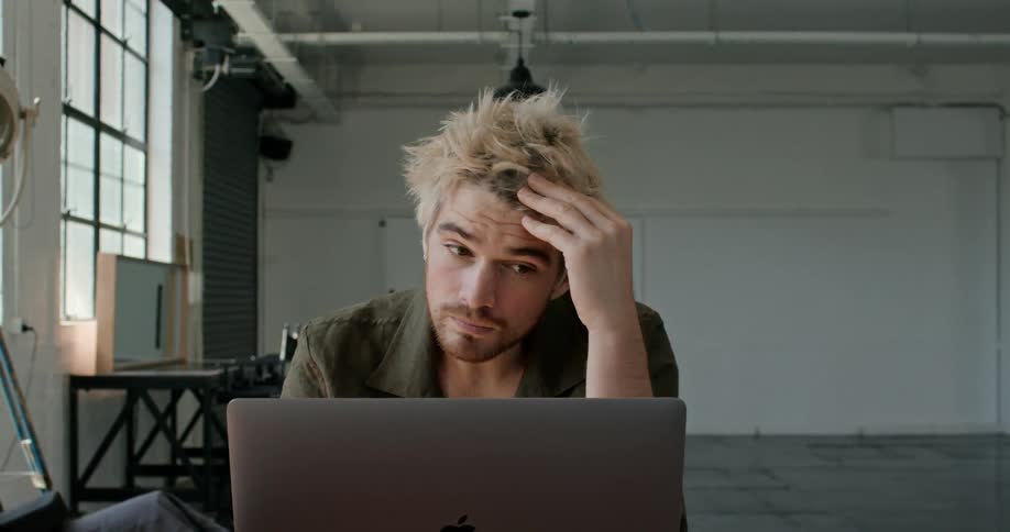 A man scratches his head and runs his hand through his hair while sitting and using a laptop