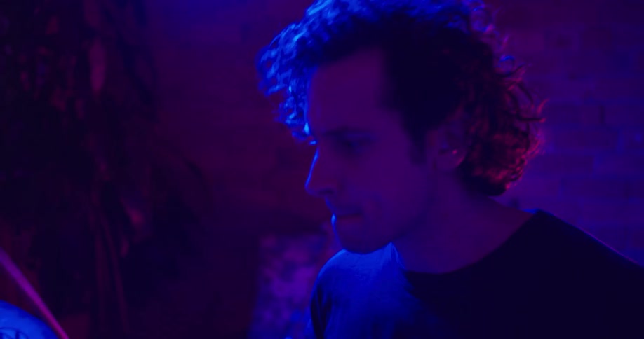 A young white man plays drums while bathed in a dark light