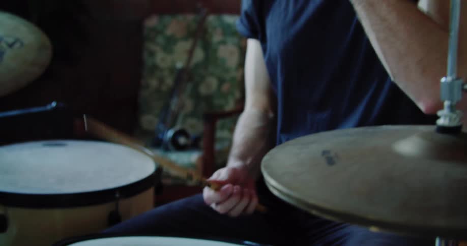 A man sits down on a stool and picks up drum sticks to start playing a drum set