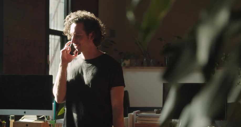 A man with curly hair stands in an office space while talking on his cell phone
