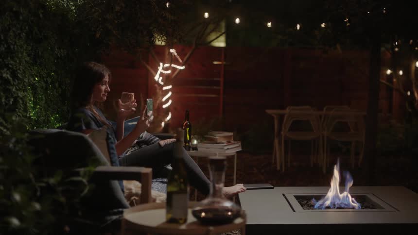 A woman sits outdoors near a fire pit while drinking wine having a video chat on her smart phone