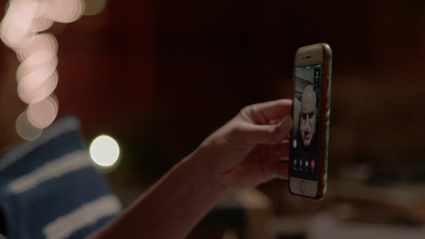 A womans hand holding up a smartphone showing the face of the man she is having a video chat with