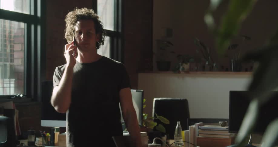 A man with curly hair stands in an office space while talking on a cell phone