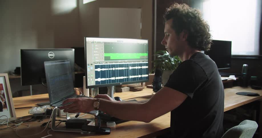 A man with curly hair sits at a desk where he is editing music using a computer