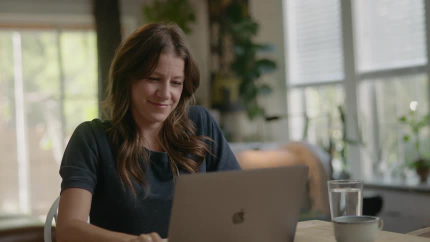 A woman smiles as she sits at a table and opens her laptop