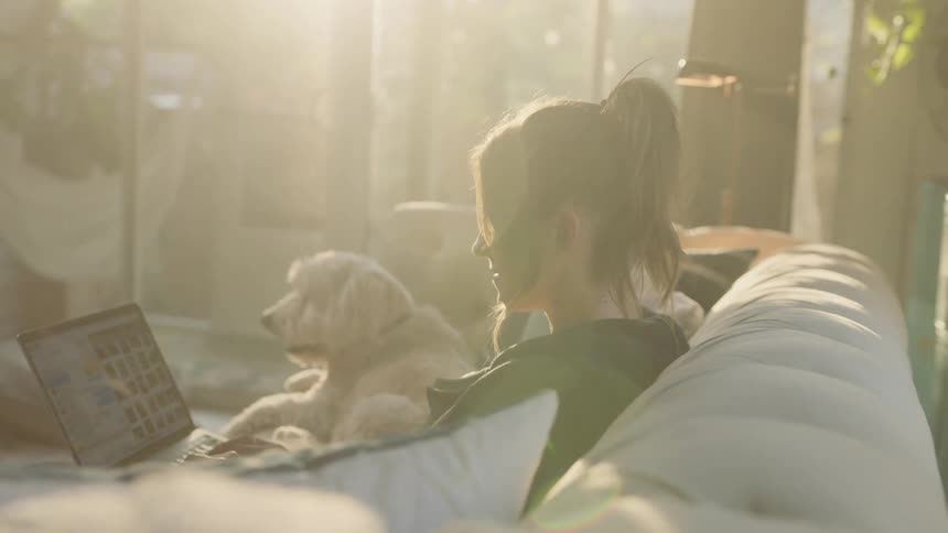 A woman sitting on a couch using a laptop next to a dog