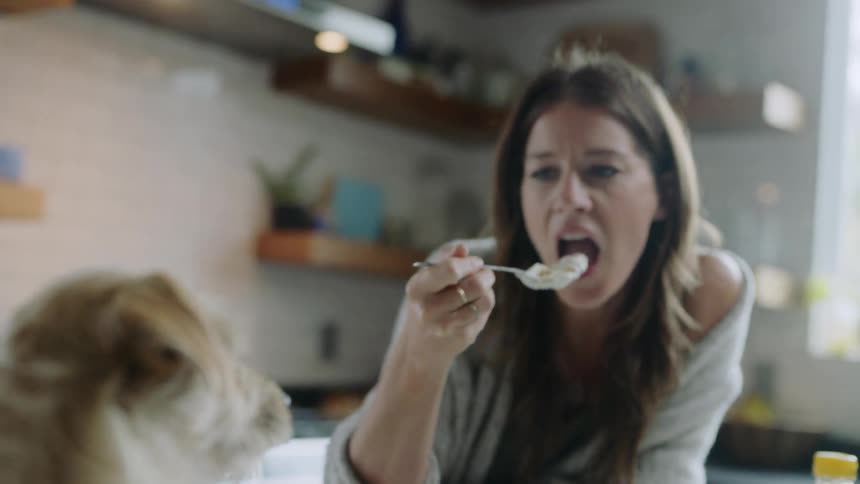 A woman sits at a table in her kitchen eating food while talking to her pet dog
