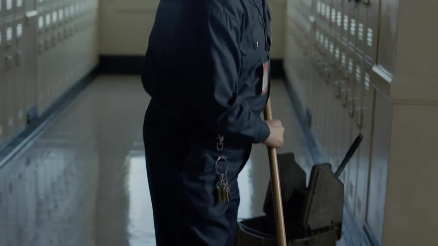 Male custodian pushes a mop on the linoleum floor of a school hallway near a set of lockers