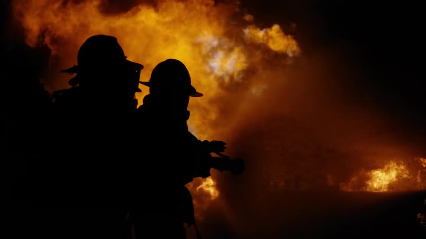Three fire fighters stand close together as one holds the nozzle of a hose to spray water onto a fire