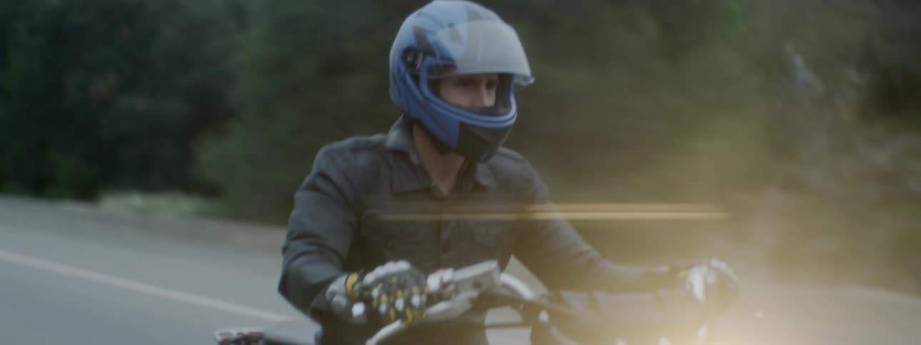 Man wearing blue helmet rides motorcycle along highway bordered by woods