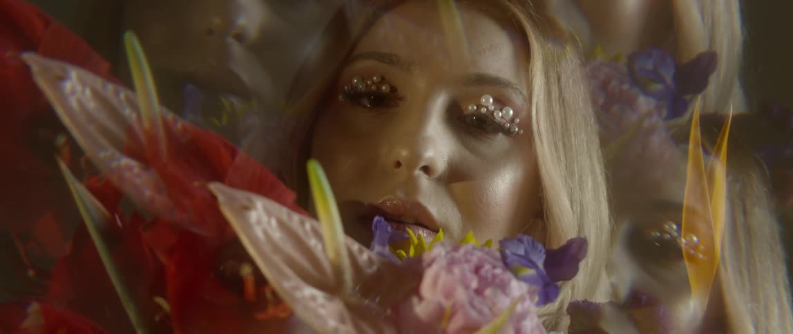 A woman with peals on her eyelids holds flowers up to her face and smells them