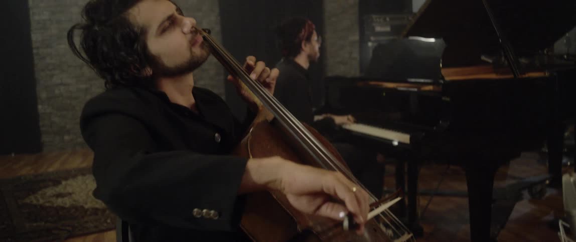 A male musician plays a cello while another plays a piano in the background